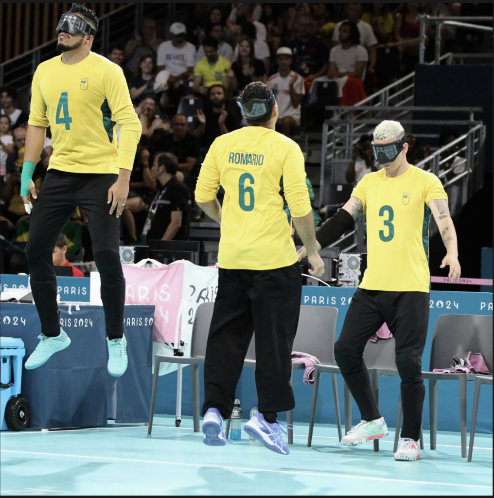 Three Brazilian athletes wearing yellow jerseys and black pants stand beside a blue court at the Paris 2024 Paralympic Games. They are wearing blindfolds or eye covers. The player on the left (number 4) is jumping slightly, while the middle player (Romario, number 6) and right player (number 3) stand on the ground. The background shows spectators in the stands and "Paris 2024" branding on barriers.